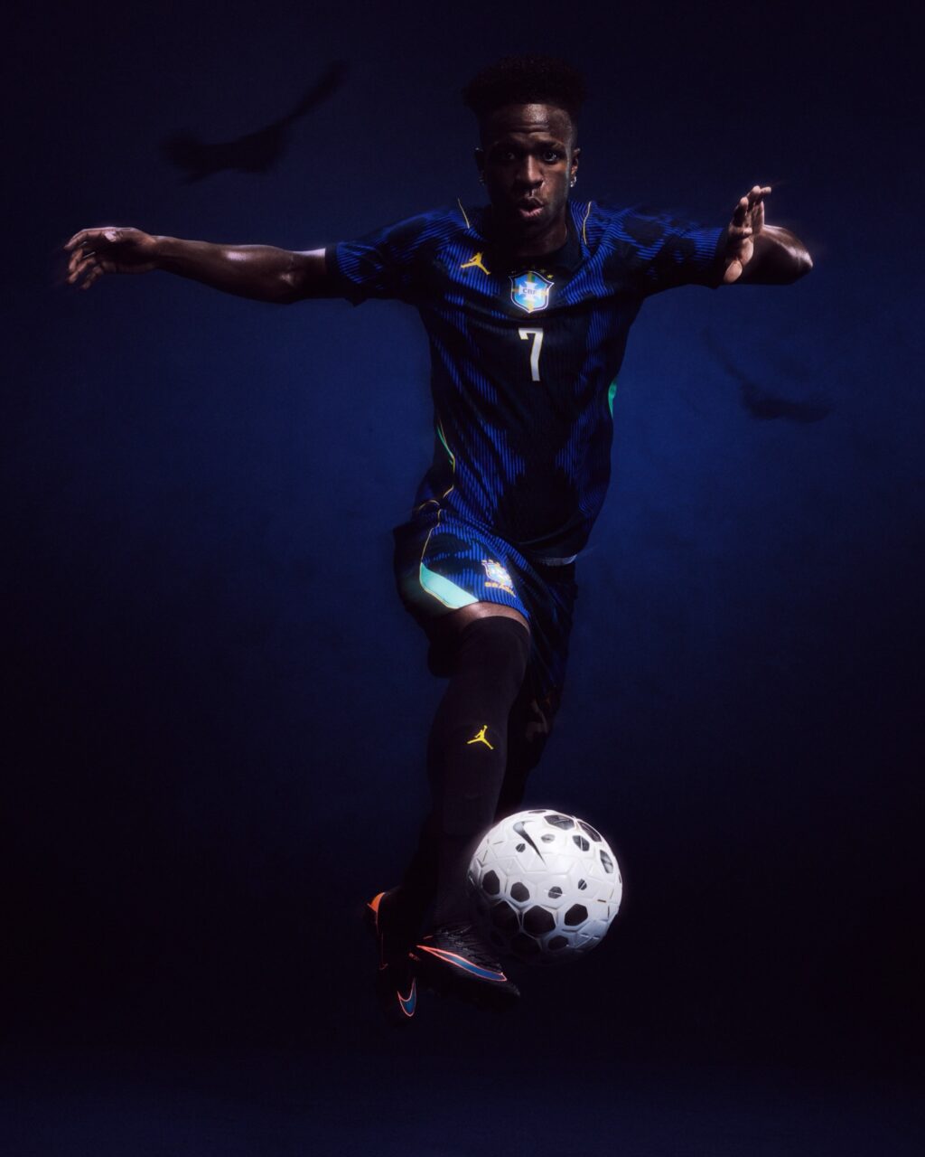 A soccer player in motion, wearing a blue and green jersey with the number 7, dribbles a soccer ball against a dark background.