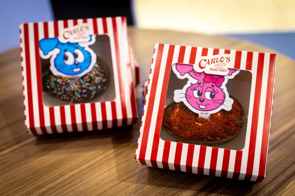Two decorated baked goods in striped takeout boxes from Carlo's Bake Shop. One features a blue bunny design, and the other has a pink bunny design.