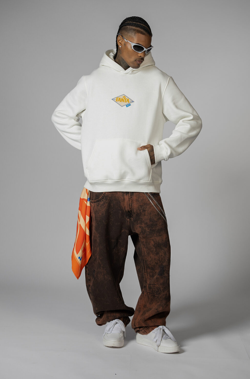 A model wearing a white hoodie with a Fanta logo, paired with brown baggy pants and white sneakers, poses confidently against a plain gray background.