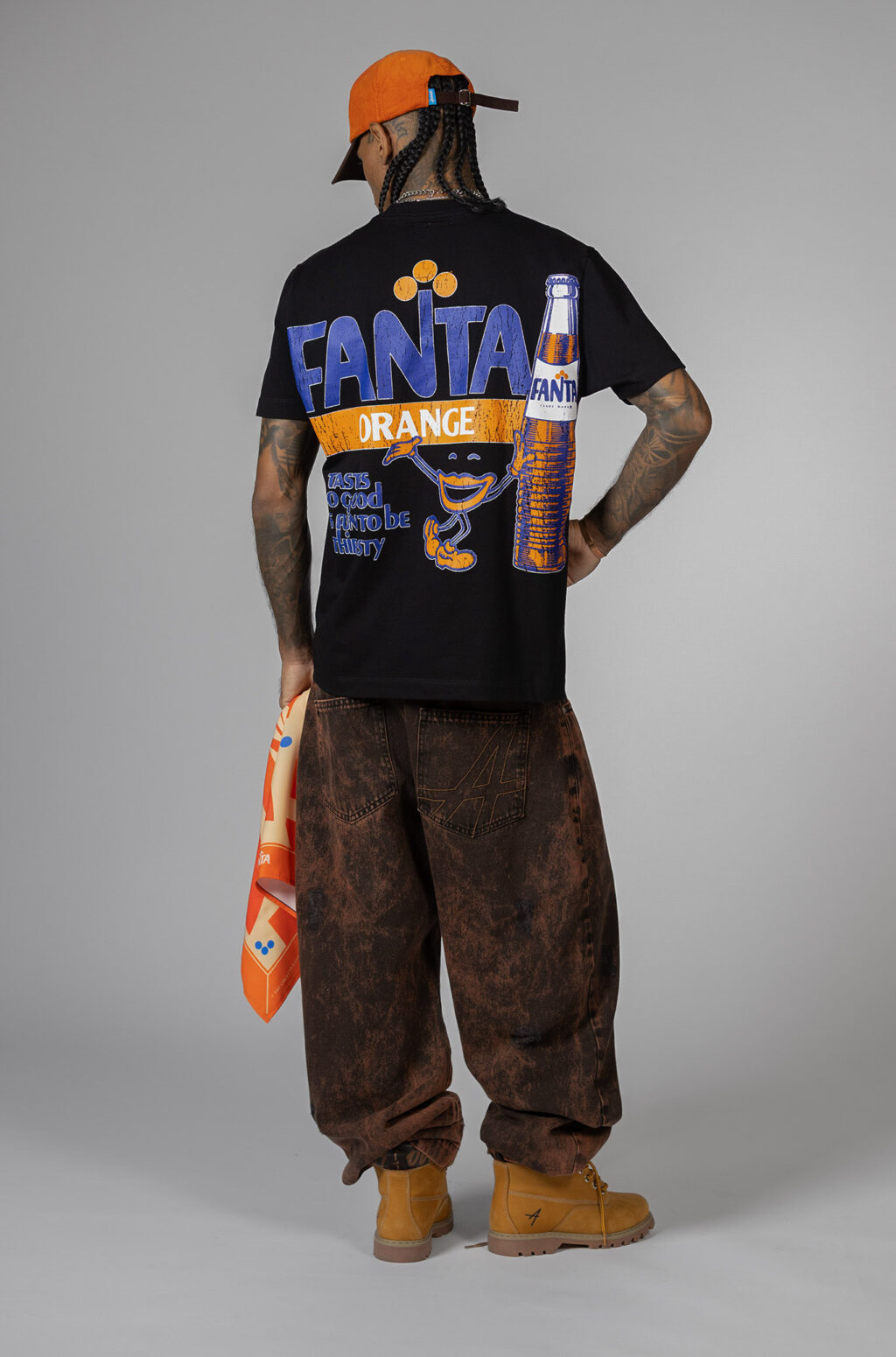 Person wearing a black Fanta-themed t-shirt with bright graphics on the back, showcasing an orange soda can character and the slogan 'Tastes so good, Fanta be thirsty.' They are also wearing brown pants and a bright orange cap.