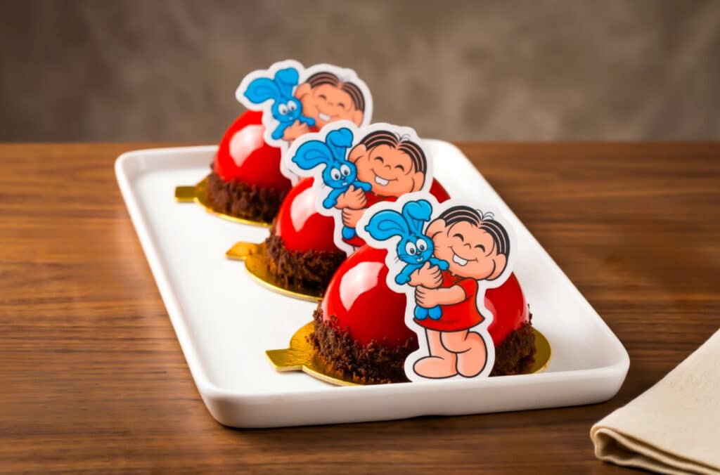 A plate with three round desserts topped with cartoon character decorations, featuring a boy hugging a blue bunny. The desserts have a glossy red coating and are placed on a wooden table.