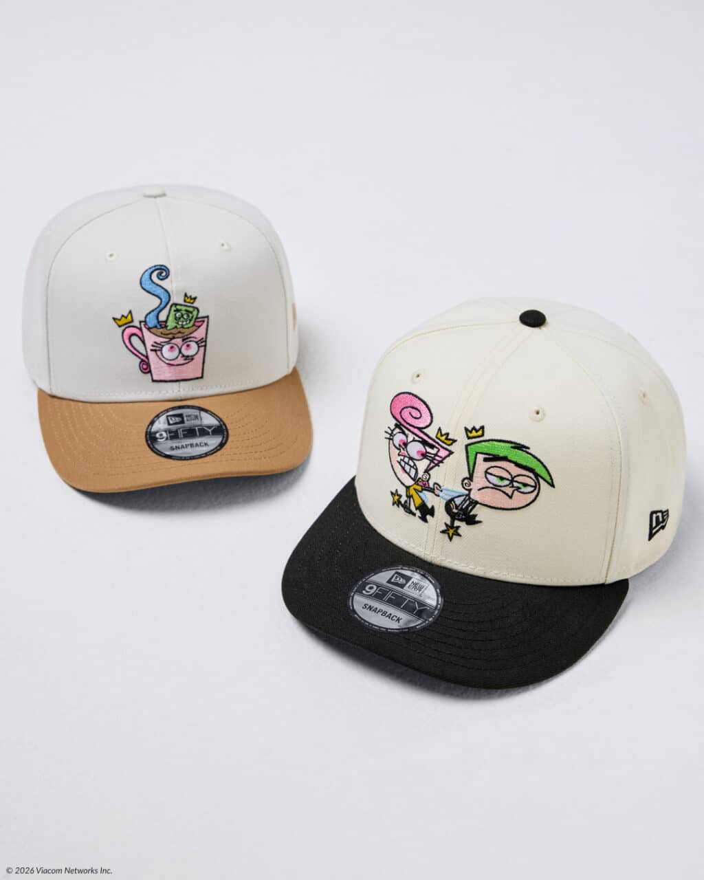 Two snapback hats featuring cartoon character designs. The left hat has a pink cup character with a green object on top, and the right hat displays two characters, one with pink hair and the other with green hair, set against a cream background and black bill.