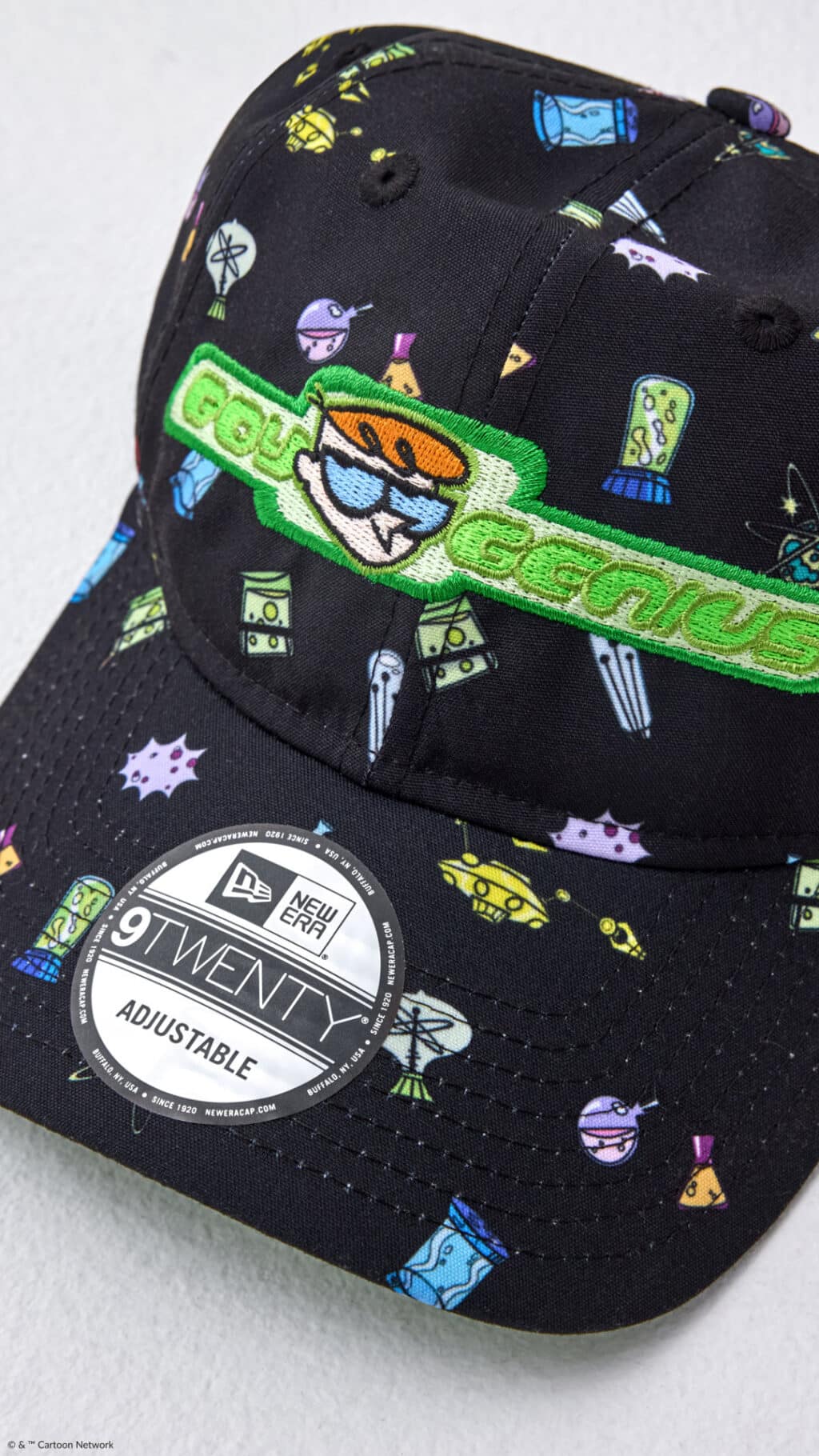 A black baseball cap featuring colorful science-themed graphics, including various scientific instruments and gadgets, with an embroidered 'Genius' logo and a character from the show Dexter's Laboratory.