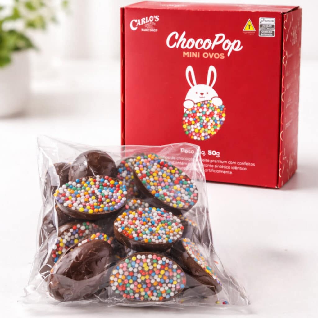 A transparent bag filled with chocolate mini eggs topped with colorful sprinkles, placed in front of a red box labeled 'ChocoPop Mini Ovos' from Carlos's Bake Shop.