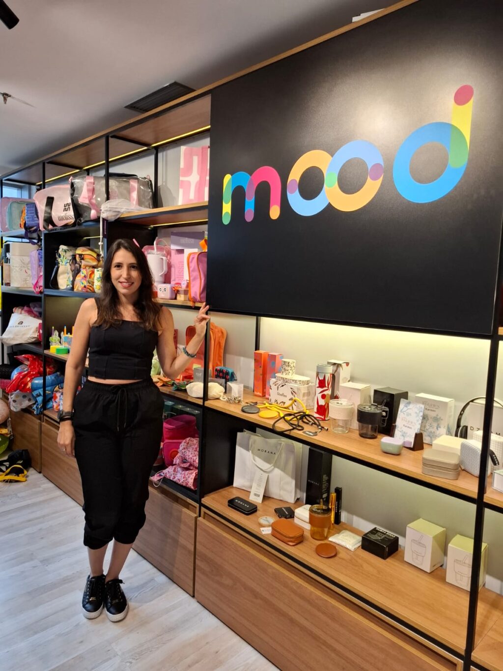 Mulher em uma loja chamada 'mood', posando ao lado de uma prateleira cheia de produtos variados, como bolsas e itens de decoração.