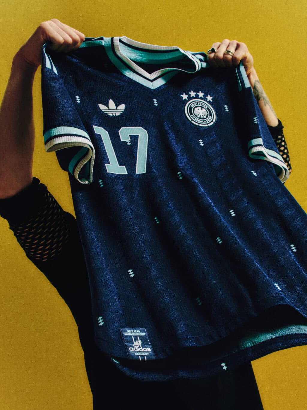 A person holding a blue soccer jersey with the number 17 and the German Football Association logo, against a bright yellow background.