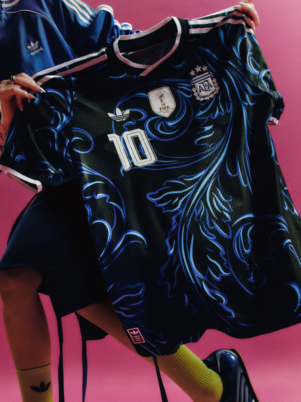 A close-up image of a green and blue Argentina soccer jersey featuring the number 10, adorned with intricate floral patterns and the Adidas logo, held against a vibrant pink background.