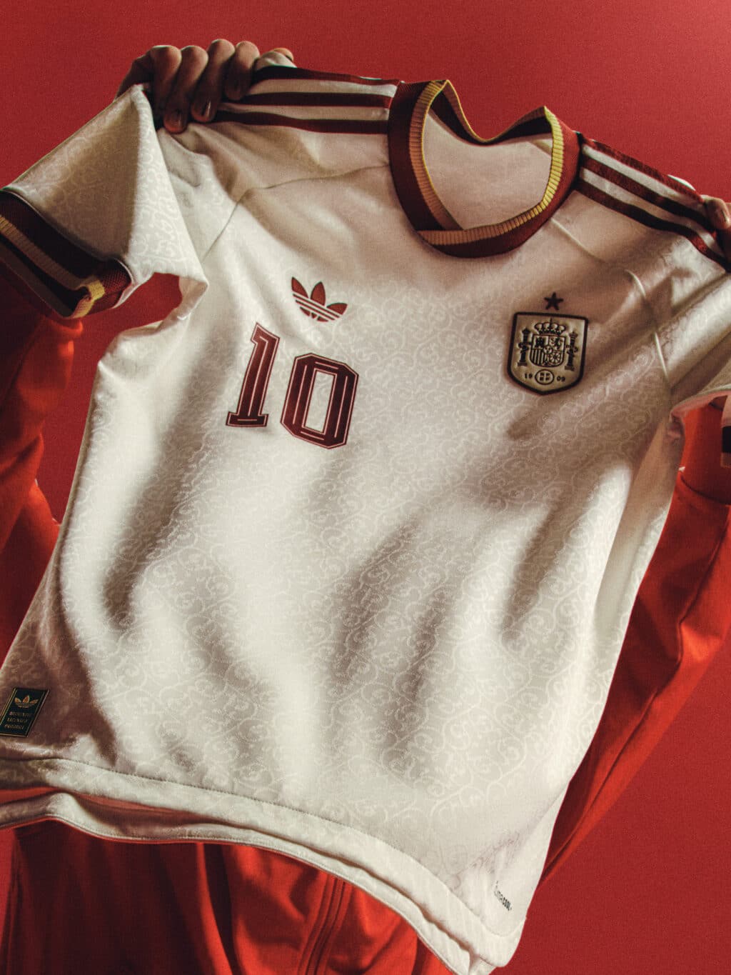 A person holding a white Spain national football jersey with red and gold accents, featuring the number 10 and an Adidas logo, against a red background.