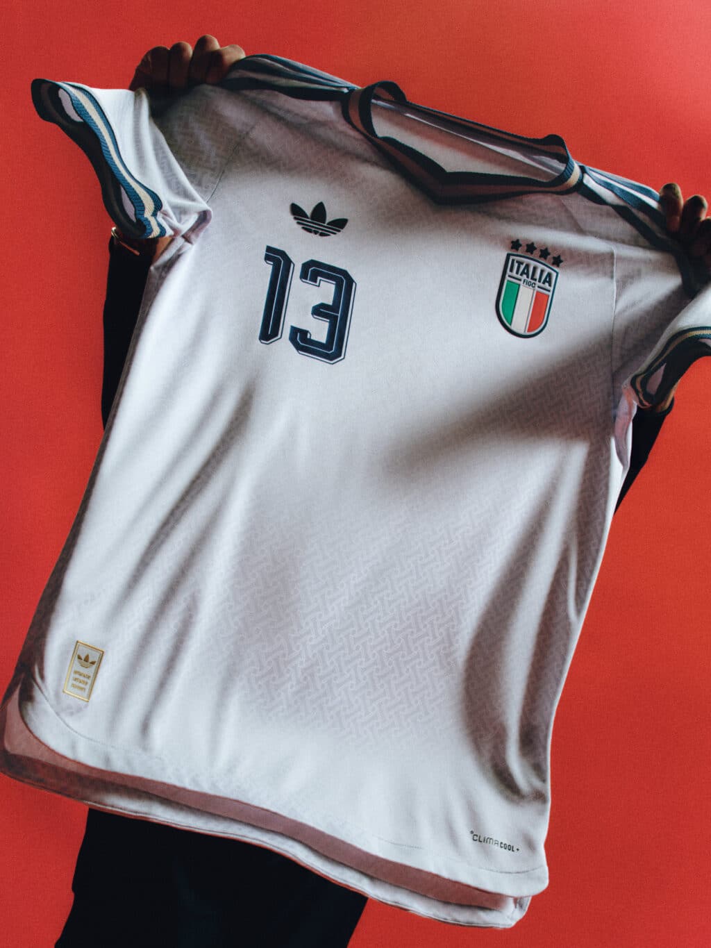 A person holding an Italian soccer jersey with the number 13, featuring the Adidas logo and the Italy national emblem, against a red background.