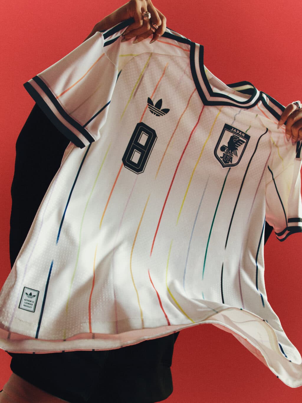 A person holding a white soccer jersey with colorful stripes, featuring the number 8 and the Japan national soccer team crest, against a red background.