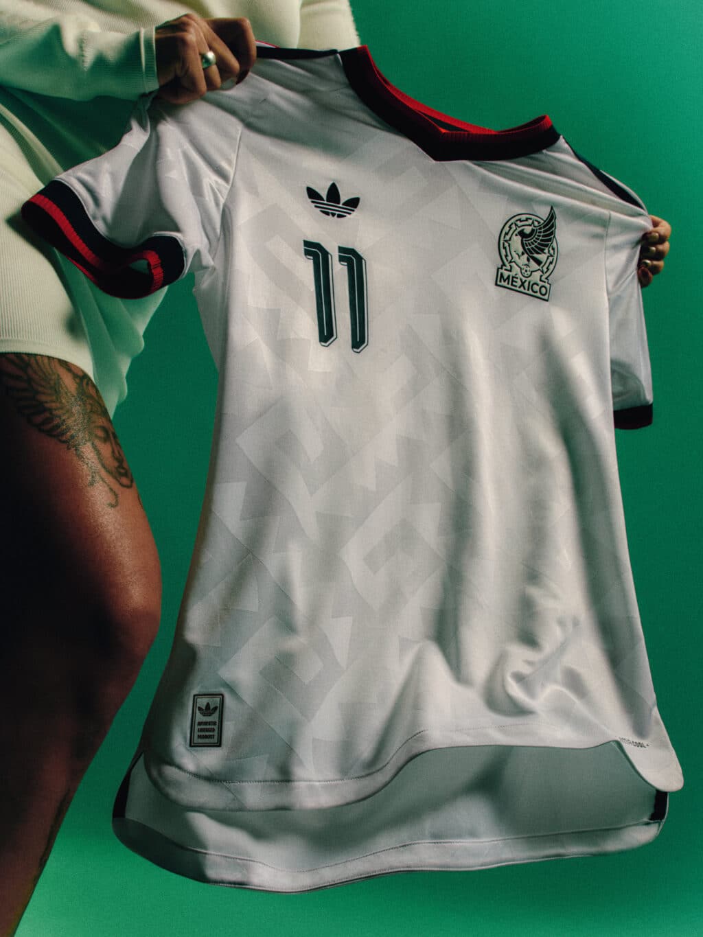 A close-up of a white soccer jersey featuring the Mexico national team logo, number 11, and the Adidas logo, held by a person against a green background.