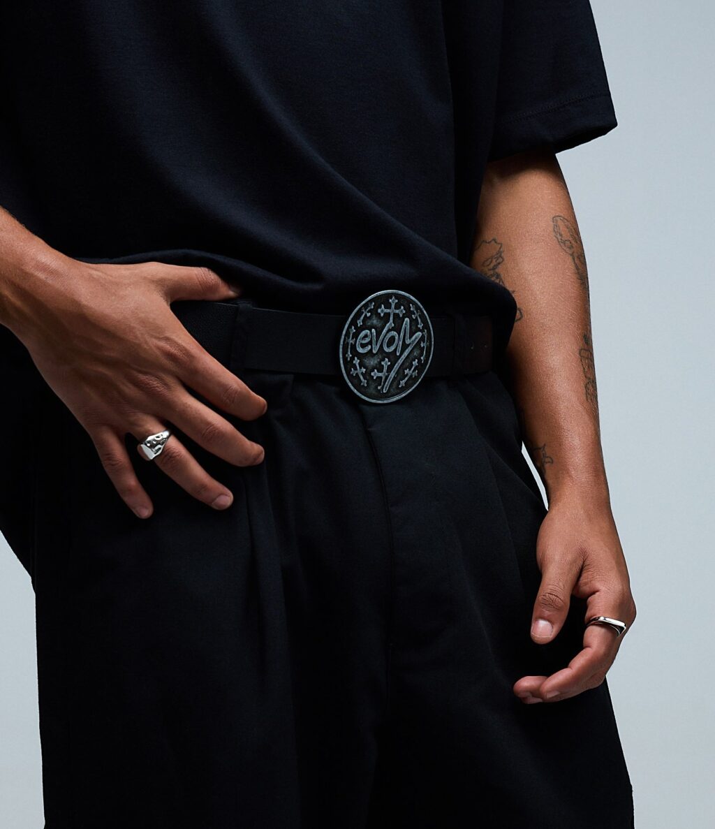 Close-up de uma pessoa vestindo camiseta preta e calça preta, mostrando uma fivela de cinto exclusiva com a palavra 'evol' e um elegante anel de prata na mão.