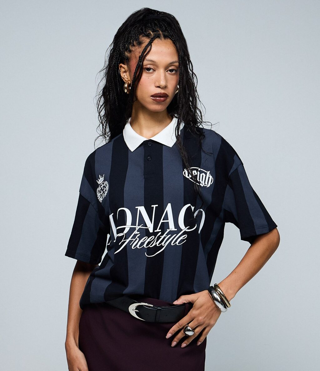 Uma modelo vestindo uma camisa polo listrada em preto e azul-marinho com a inscrição 'MONACO Freestyle', combinada com uma saia bordô. A modelo tem cabelos longos trançados e usa joias como acessórios.