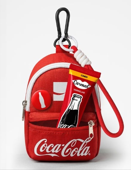 Uma pequena mochila vermelha com a marca Coca-Cola, com um zíper que, ao ser aberto, revela um tubo de condimento vermelho e um emblema circular.