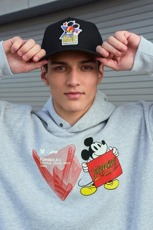 A young man wearing a gray hoodie featuring Mickey Mouse and a graphic design, holding a red sign. He is also wearing a black cap with a colorful logo.