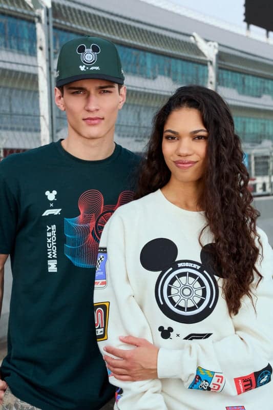 A young man wearing a black t-shirt with a Mickey Mouse graphic and a green cap stands next to a young woman. She is wearing a white sweatshirt featuring a Mickey Mouse design and colorful patches. They pose confidently in front of a racetrack backdrop.