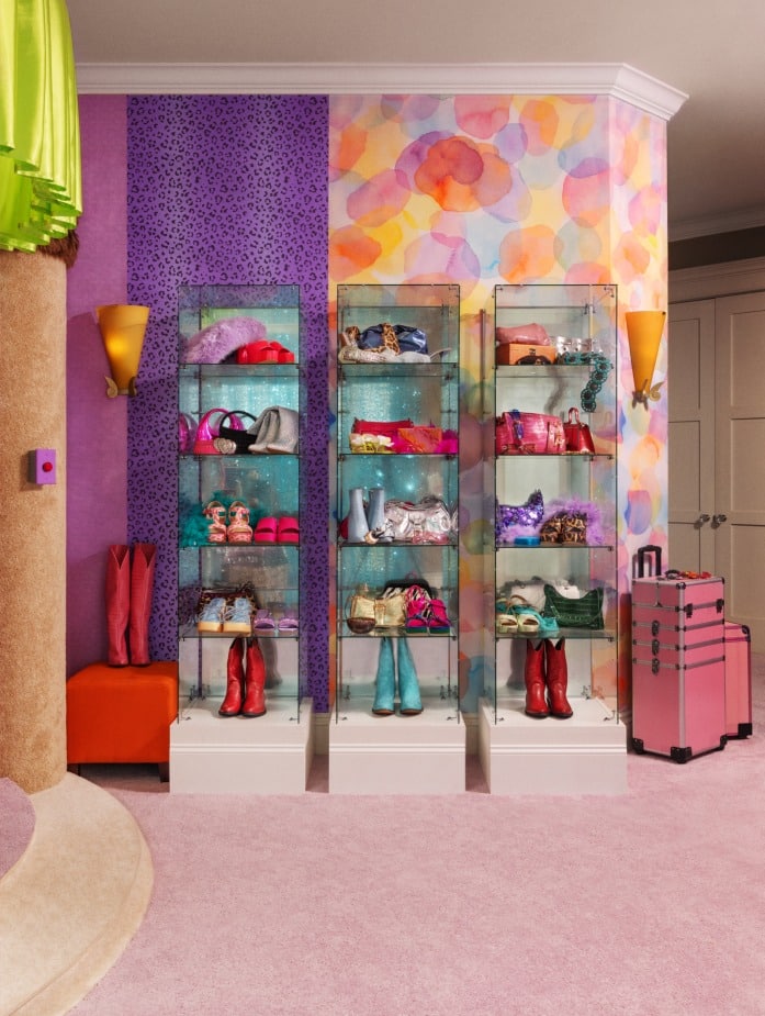 Uma vitrine colorida com calçados e bolsas estilosas, exposta em vitrines de vidro, contrasta com uma parede vibrante com estampas em tons de roxo e pastel. O ambiente conta com carpete macio e detalhes decorativos.