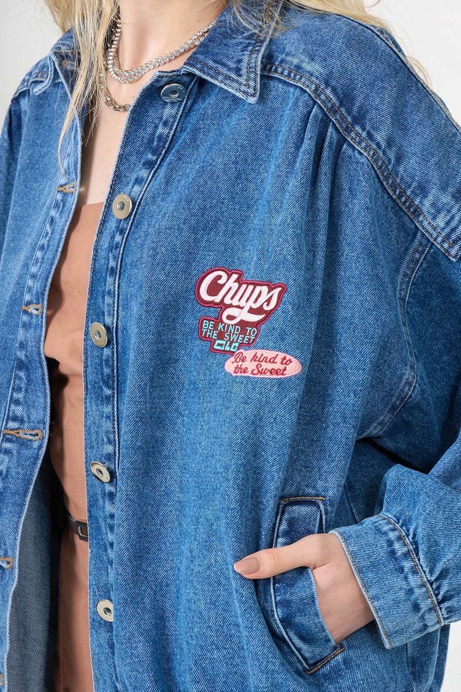 Uma pessoa vestindo uma jaqueta jeans azul com o logotipo "Chups" bordado e a frase "Seja gentil com os doces". A jaqueta tem um corte folgado e é combinada com uma blusa marrom.