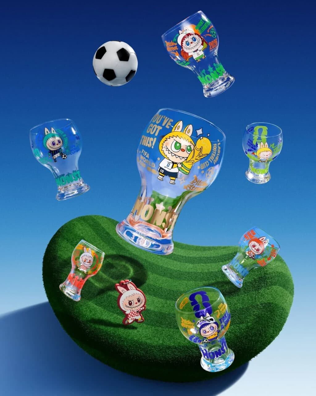 Copos de vidro coloridos com personagens de desenhos animados e temas da Copa do Mundo da FIFA, dispostos sobre uma superfície gramada verde com uma bola de futebol ao fundo.