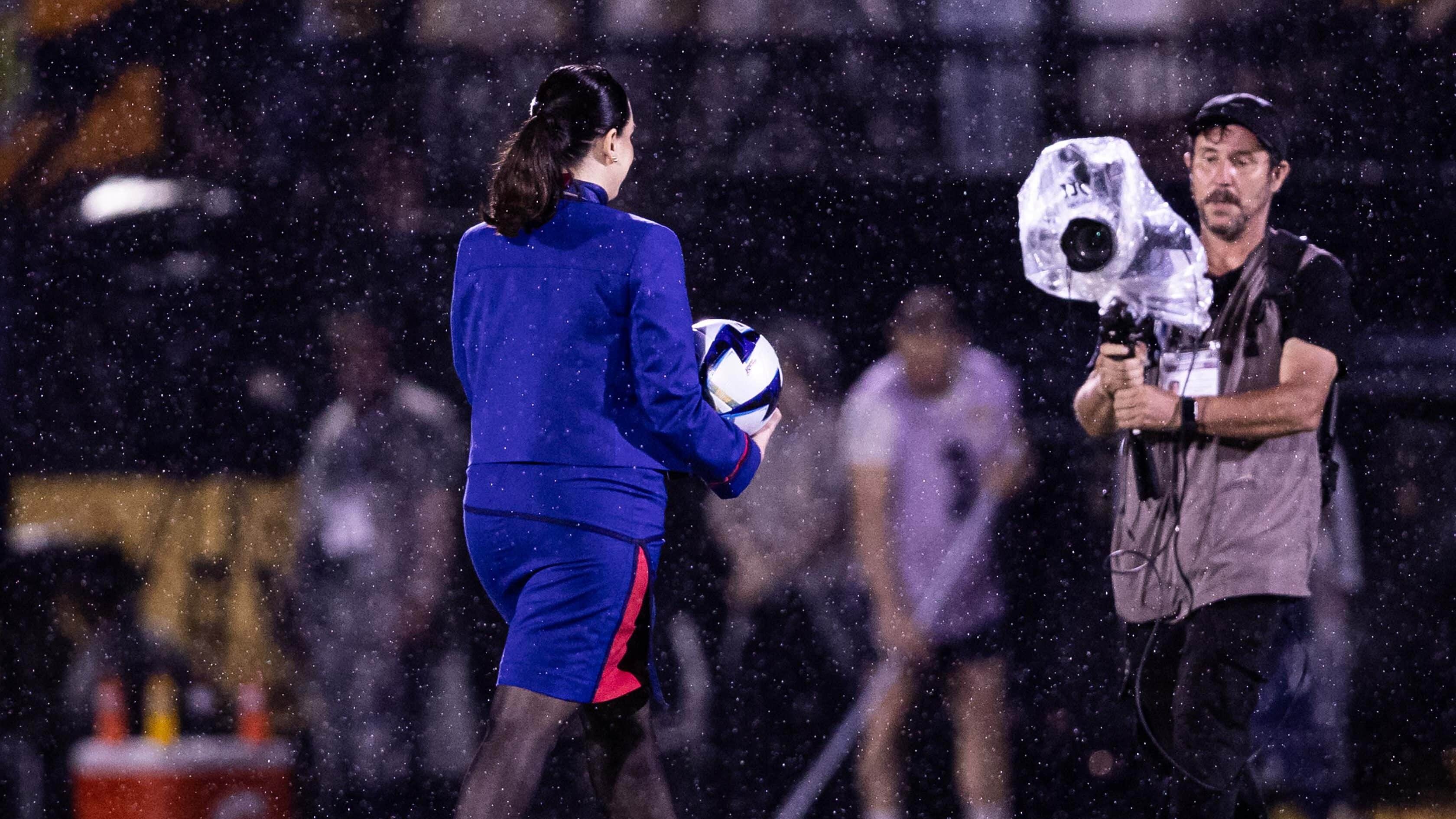 Uma mulher de terno azul, segurando uma bola de futebol, caminha por um campo encharcado pela chuva enquanto um homem com uma câmera se prepara para registrar o momento.