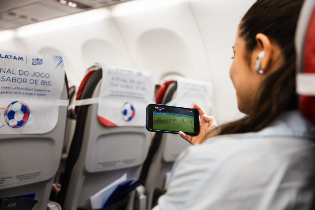 Uma mulher sentada em um avião assistindo a um jogo esportivo em seu smartphone com fones de ouvido, enquanto as poltronas do avião são visíveis ao fundo.