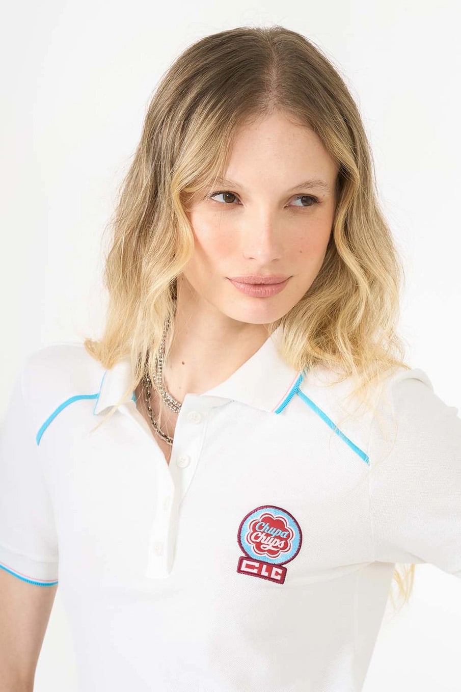 Uma mulher com cabelos loiros ondulados vestindo uma camisa polo branca com detalhes em azul e o logotipo da Chupa Chups.