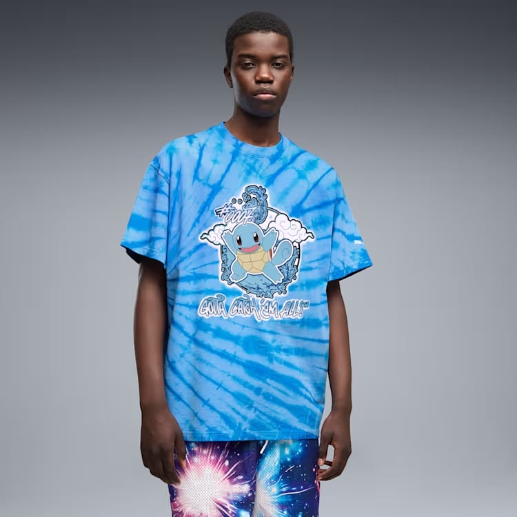 Uma pessoa vestindo uma camiseta azul tie-dye com um personagem de desenho animado e a frase "Gotta Catch 'Em All!" (Temos que pegar todos!), ao lado de shorts estampados e vibrantes.
