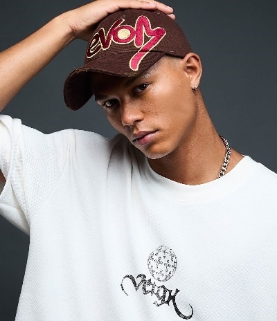 Um jovem de cabelo curto, usando um boné marrom com a palavra "evom" e uma camiseta branca larga com estampa. Ele está posando contra um fundo escuro.
