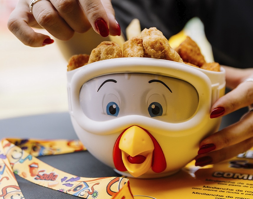 Uma tigela em formato de cabeça de galinha cheia de nuggets de frango, com uma mão humana estendida para pegar alguns.