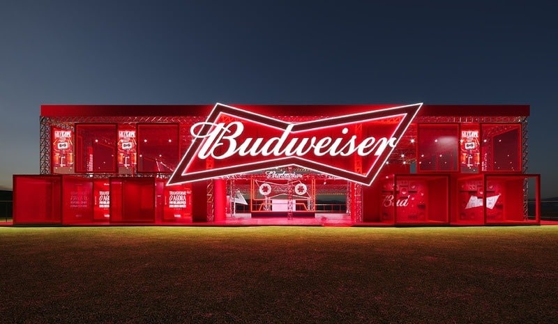 Uma grande estrutura iluminada com o logotipo da Budweiser em destaque, apresentando um esquema de cores vermelho contra um fundo crepuscular.