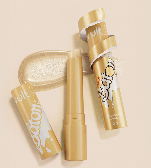Um batom líquido dourado em formato de bastão, com a embalagem, disposto horizontalmente ao lado de uma amostra do produto sobre um fundo claro.