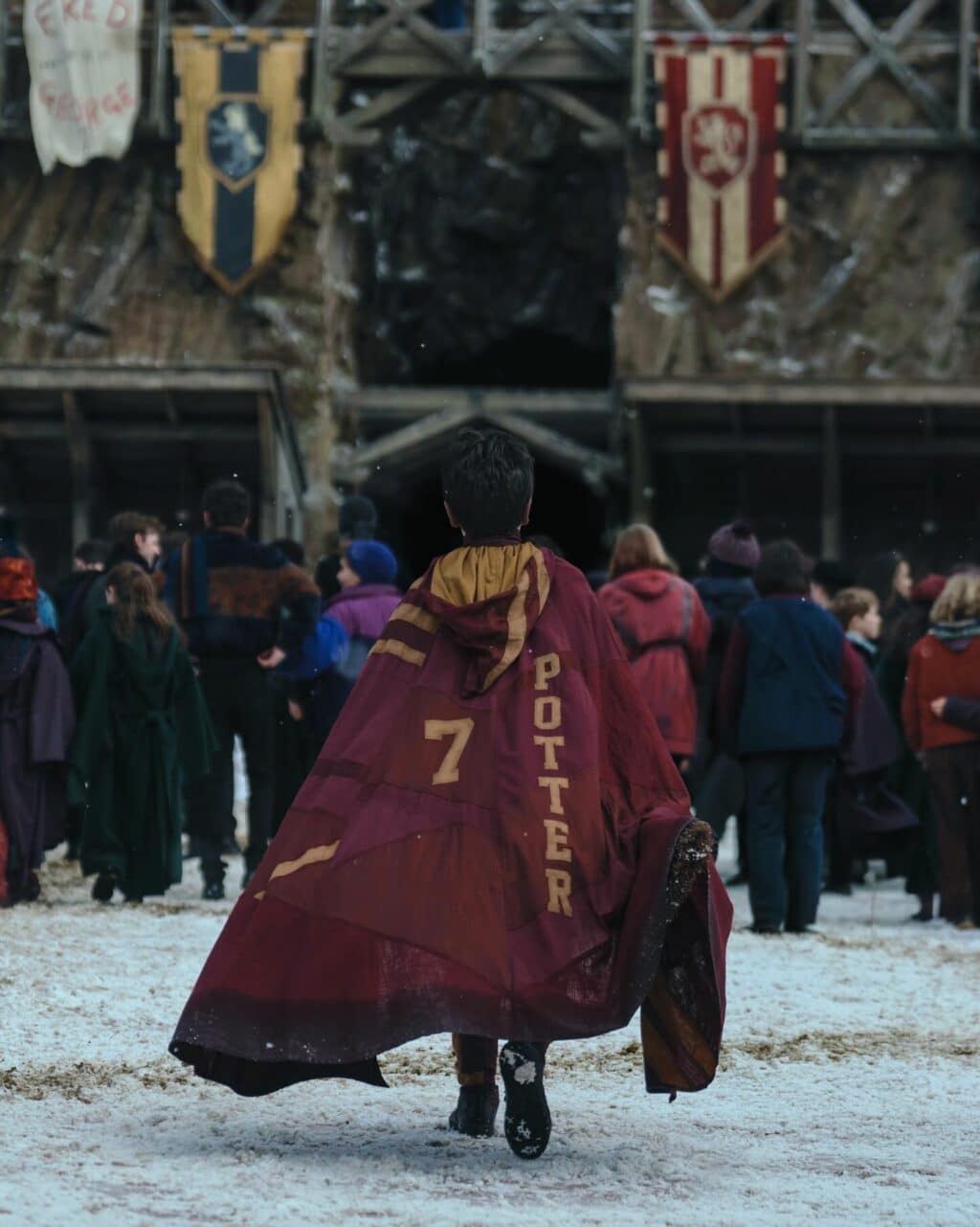 Uma criança com uma capa marrom e dourada, com o nome 'POTTER' e o número '7', caminhando em direção a um grupo de pessoas em um ambiente nevado.