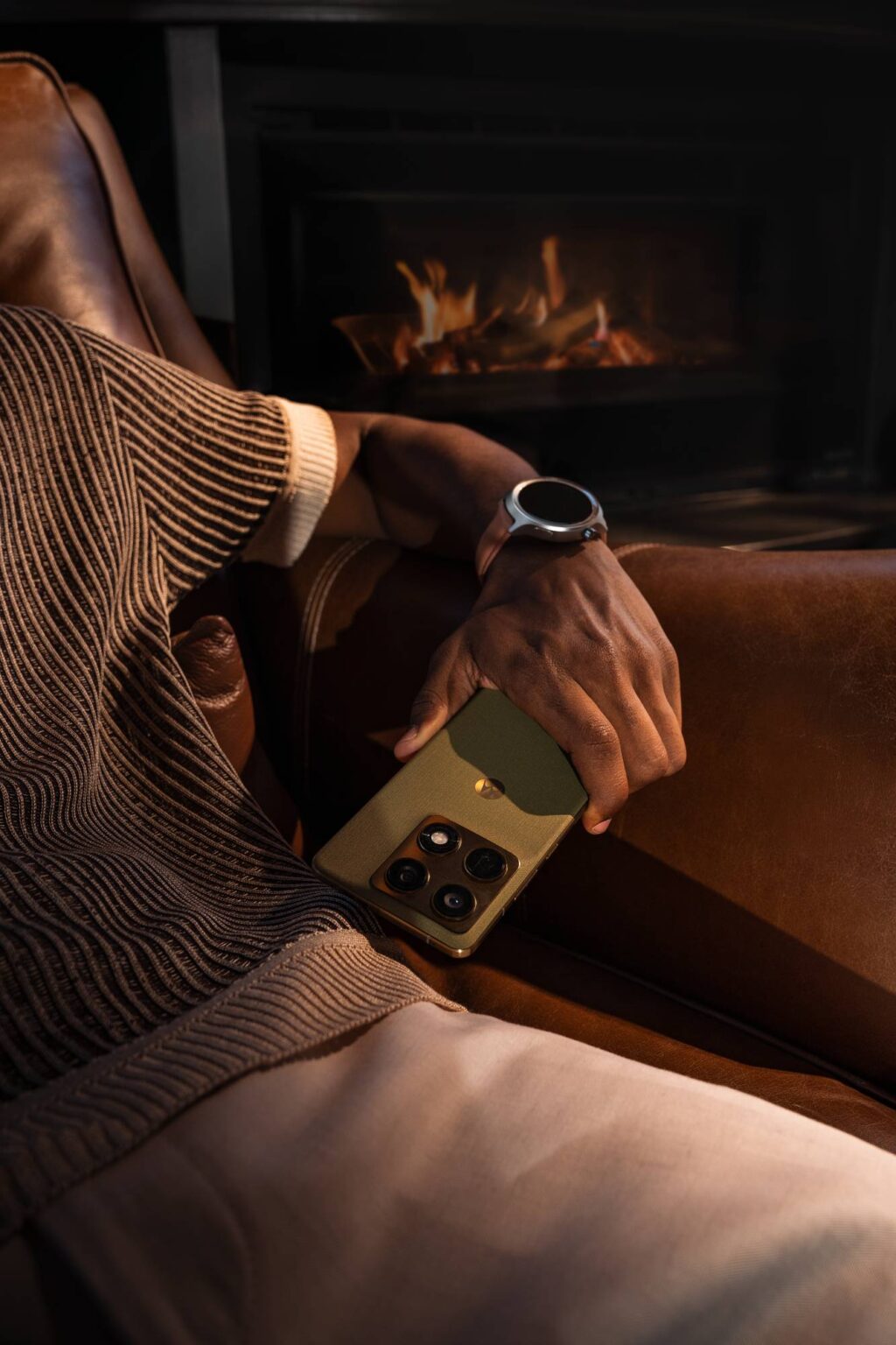 Uma mão segurando um smartphone verde com câmera enquanto está descansando em uma poltrona de couro, com uma lareira acesa ao fundo.