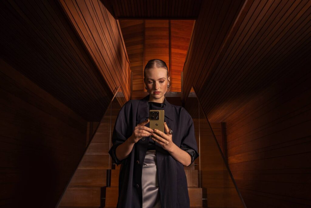 Mulher segurando um celular enquanto está em uma escada com paredes de madeira ao fundo, iluminada de forma suave.