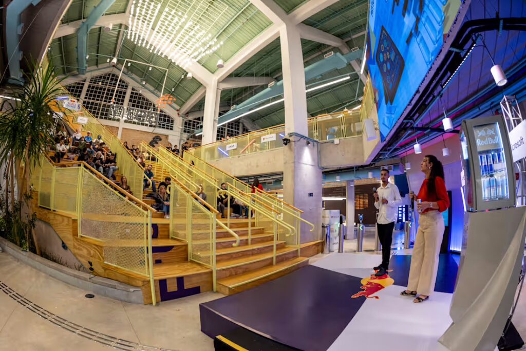 Pessoas assistindo a uma apresentação em um espaço moderno, com duas pessoas no palco, uma segurando um microfone e outra segurando uma bebida, e uma tela grande ao fundo.
