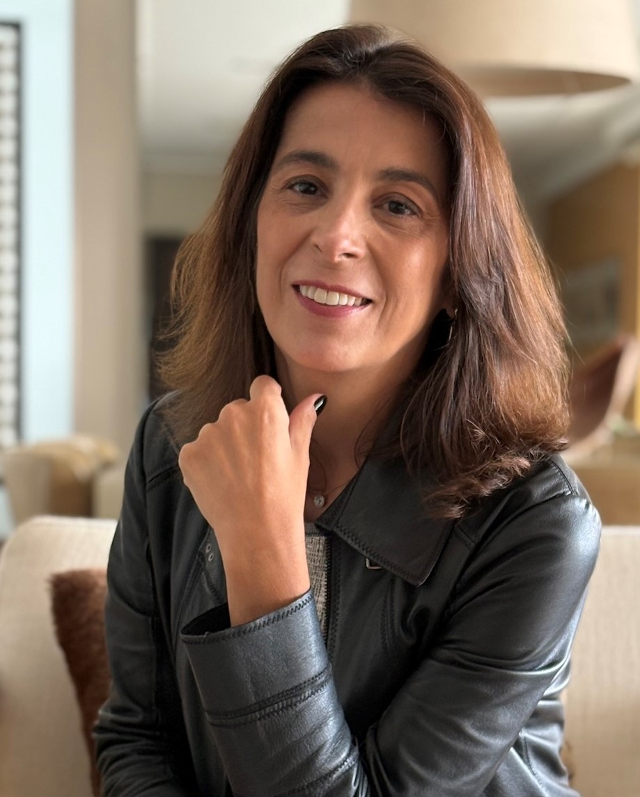 Mulher sorridente com cabelo castanho claro, usando uma jaqueta de couro preta e um suéter claro, posando em um ambiente interno iluminado.