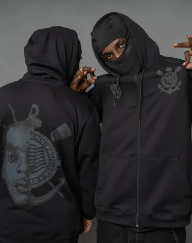 Two individuals wearing black hoodies, one displaying a graphic of a face and logo on the back, while both have hoods up and one is partially covering their face with a mask.