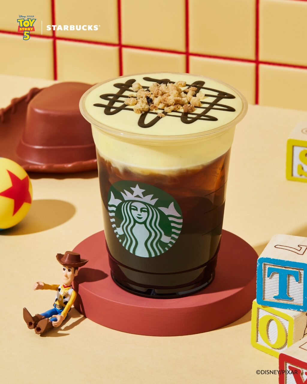 A Starbucks drink featuring a layered coffee with a creamy top, decorated with chocolate drizzle and cookie crumbs, placed on a circular red platform. A small toy figure of Woody from Toy Story sits beside the drink, with colorful building blocks in the scene.