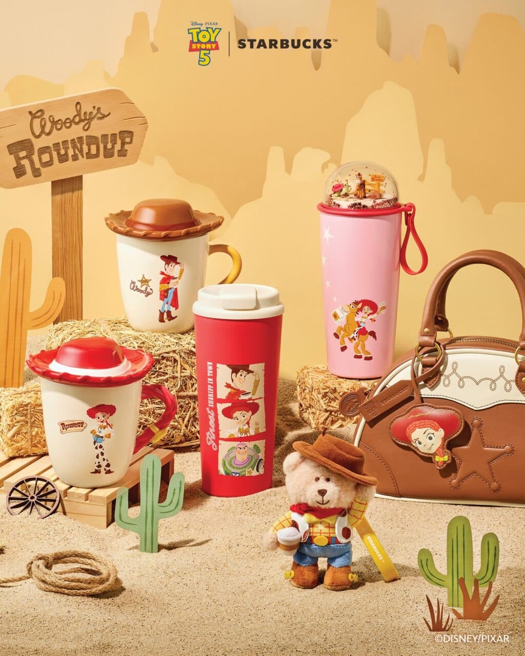 A display featuring various Toy Story 4 themed Starbucks mugs and cups alongside a plush bear dressed as Woody. The setting includes wooden and cactus decorations with a sign saying 'Woody's Roundup'.