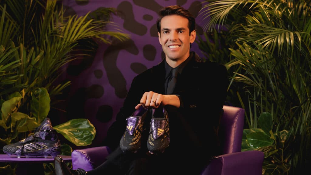Homem segurando chuteiras Adidas Predator Kaká Signature em cenário com folhagens e fundo roxo