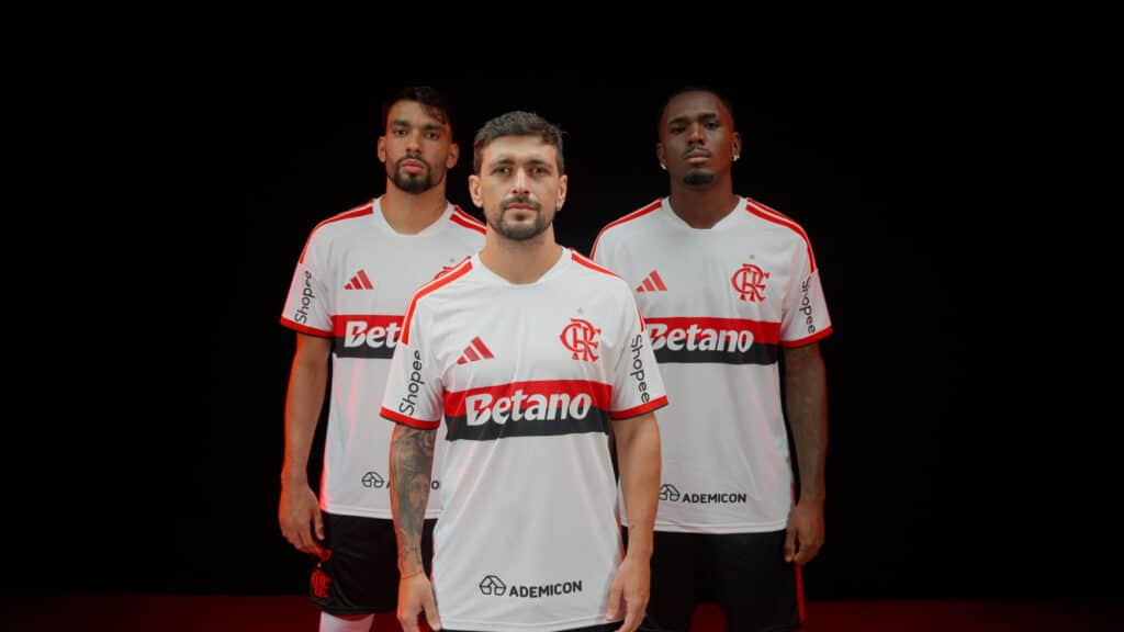 Três jogadores do Flamengo posam em uniforme branco e vermelho, com fundo escuro e logotipos do clube.