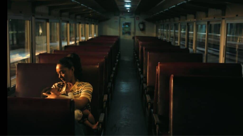 Interior de um trem à noite, com uma mulher sentada em um banco e o corredor vazio ao fundo.