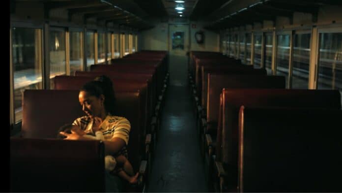 Interior de um trem à noite, com uma mulher sentada em um banco e o corredor vazio ao fundo.