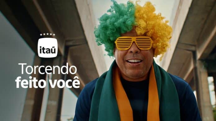 Ronaldo Fenomeno sorrindo com óculos listrados de cores verde e amarelo, em campanha do Itaú Torcendo feito você.