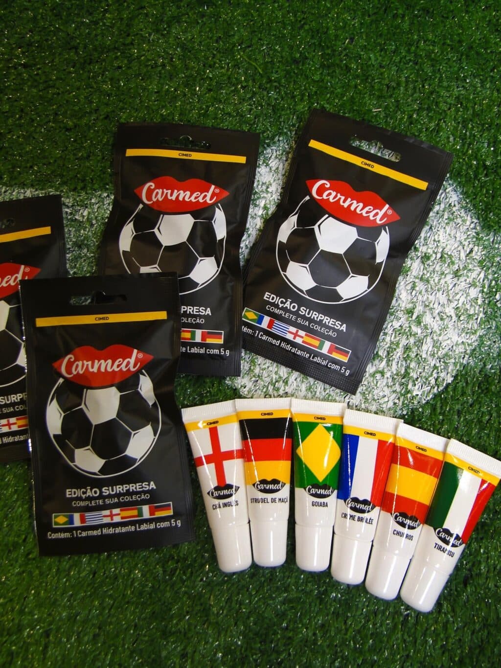 Hidratantes labiais Carmed com tema de futebol, em embalagens e cartela sobre gramado verde.