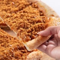 Pizza doce com cobertura de biscoito Biscoff em pedaço sendo puxado à mão, com calda e migalhas sobre a massa.