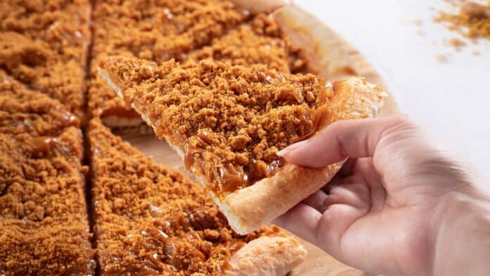 Pizza doce com cobertura de biscoito Biscoff em pedaço sendo puxado à mão, com calda e migalhas sobre a massa.
