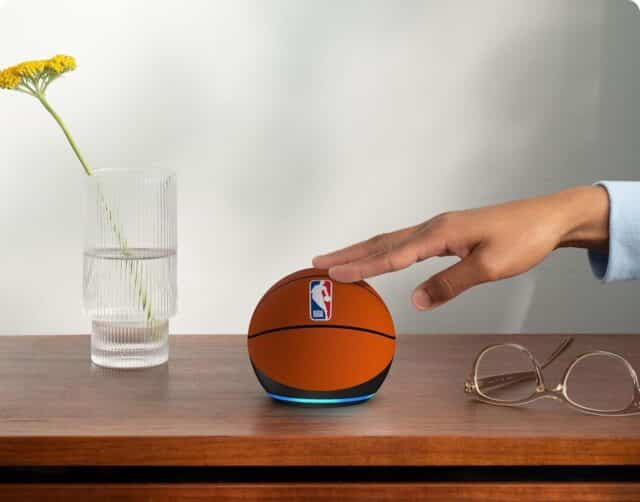 Pessoa tocando no Echo Dot edição NBA em cima de uma mesa, com vaso e óculos ao lado