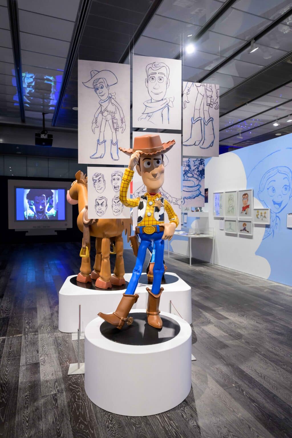 Uma estatueta em tamanho real do Woody, o personagem cowboy de Toy Story, em um pedestal em uma exposição, ao lado de uma figura de cavalo e esboços do Woody ao fundo.