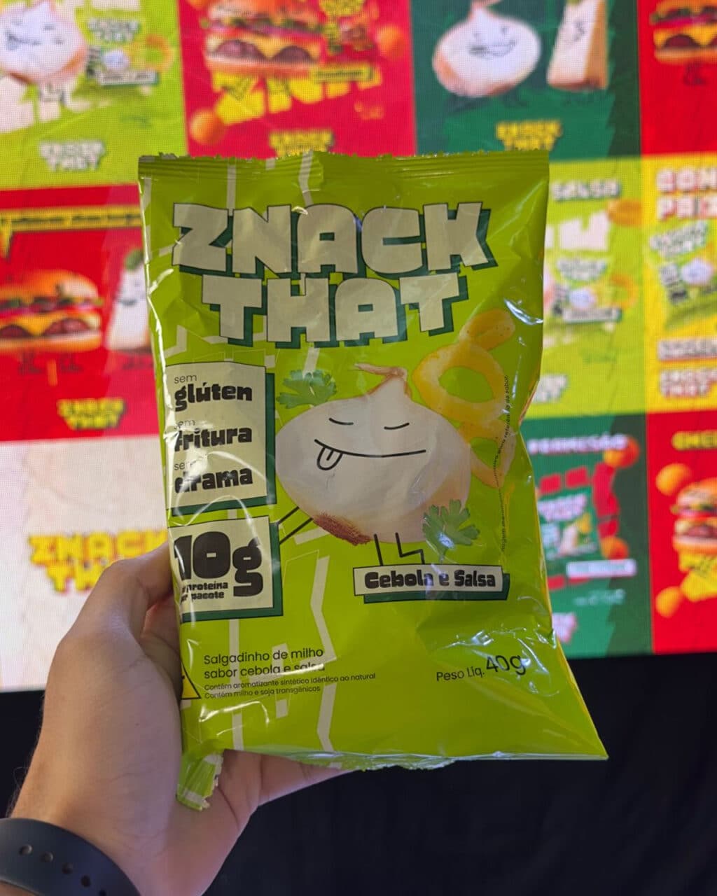 Uma mão segurando uma embalagem verde brilhante do salgadinho de milho 'ZNAK THAT', com um personagem de cebola de desenho animado e o sabor 'Cebolla e Salsa' (Cebola e Salsa). Ao fundo, imagens promocionais coloridas de diversos salgadinhos são exibidas.